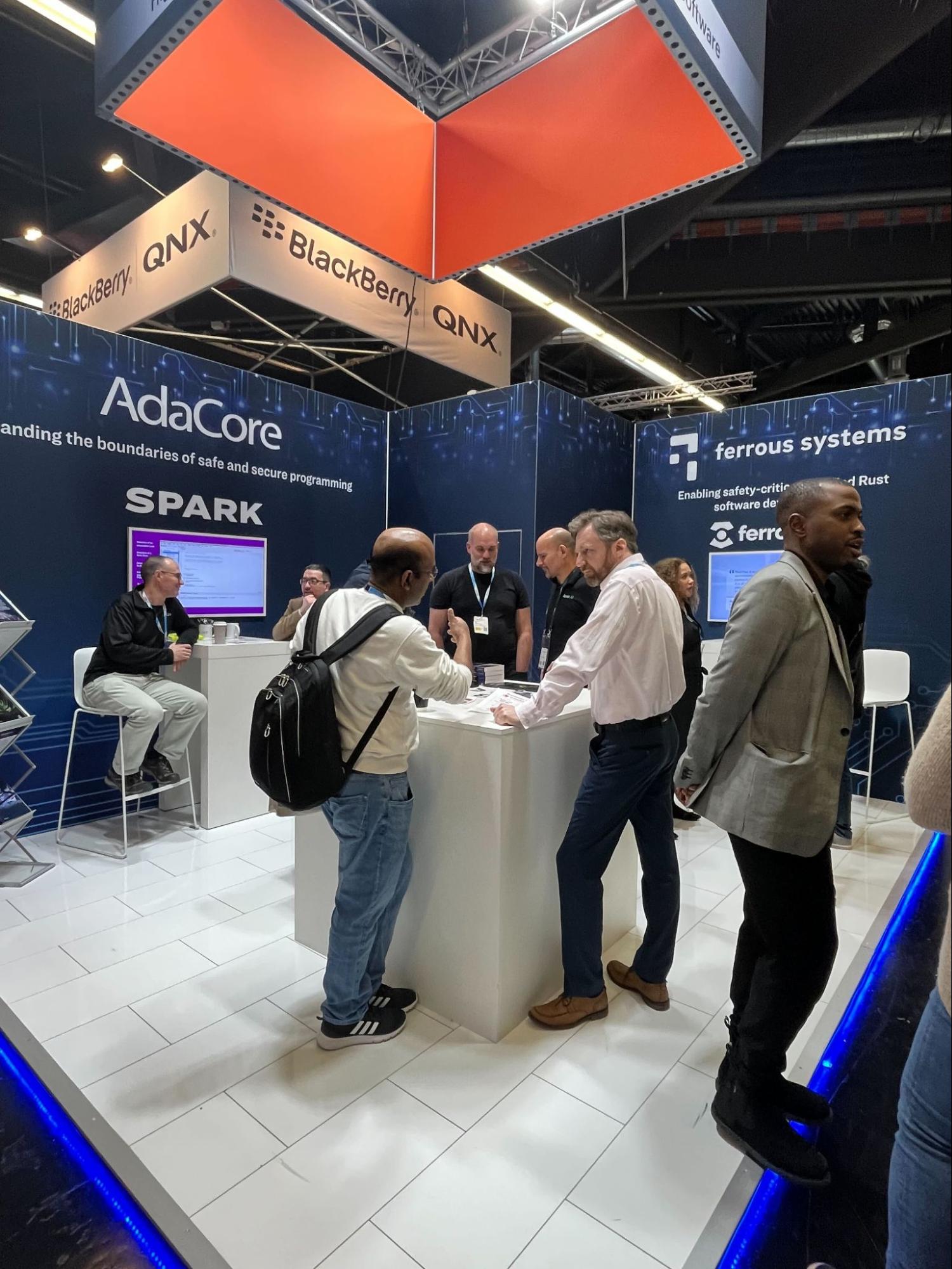 Several people standing around a central table in a booth. The left side promotes Ada/SPARK, the right side promotes Ferrocene/Rust